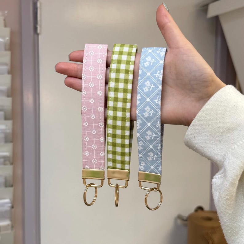 May include: Three fabric keychains in pink floral, green gingham, and blue floral patterns. Each keychain has a gold-colored metal clasp and ring. The keychains are held up in a hand.