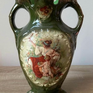 Op de afbeelding: Groene keramische vaas met twee handvatten en een decoratief bloemmotief. De vaas heeft een geschilderde scène van een vrouw en een kind in een tuin. De rand en de handvatten hebben een goudkleurige rand.