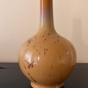 Puede incluir: Un jarrón de cerámica con cuerpo bulboso y cuello largo y delgado. El jarrón tiene un degradado de color, pasando de un beige claro en la base a un naranja más oscuro en la parte superior. Está adornado con marcas abstractas de color marrón oscuro.