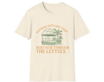 Puede incluir: Camiseta color crema con un gráfico verde y naranja. El gráfico presenta un barco, palmeras y el texto "NOTHING HITS LIKE THAT BOAT RIDE THROUGH THE LETTUCE". La camiseta tiene cuello redondo y mangas cortas.