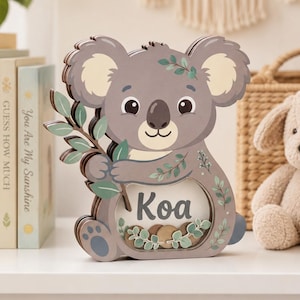 Può includere: Un salvadanaio in legno a strati a forma di koala con un pannello frontale trasparente, il nome Koa stampato in grigio scuro e illustrazioni decorative di foglie di eucalipto verdi. Il koala ha il pelo grigio, le orecchie color crema e tiene un ramo.