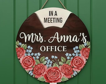 May include: Round wooden office sign with a dark brown background and a floral border of red roses and blue flowers. The sign reads "IN A MEETING" at the top and "Mrs. Anna's OFFICE" in white script.