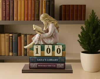 May include: A figurine of a woman reading a book, seated atop a stack of books. The books display the text "SARA'S LIBRARY" and "BOOKS READ THIS YEAR". The stack of books has the number "100" on top. A small evergreen tree in a white pot is to the right.