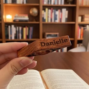 May include: A wooden bookmark with the name "Danielle" engraved on it. The bookmark is held above an open book, with a bookshelf in the background. The bookmark is made of a light brown wood.