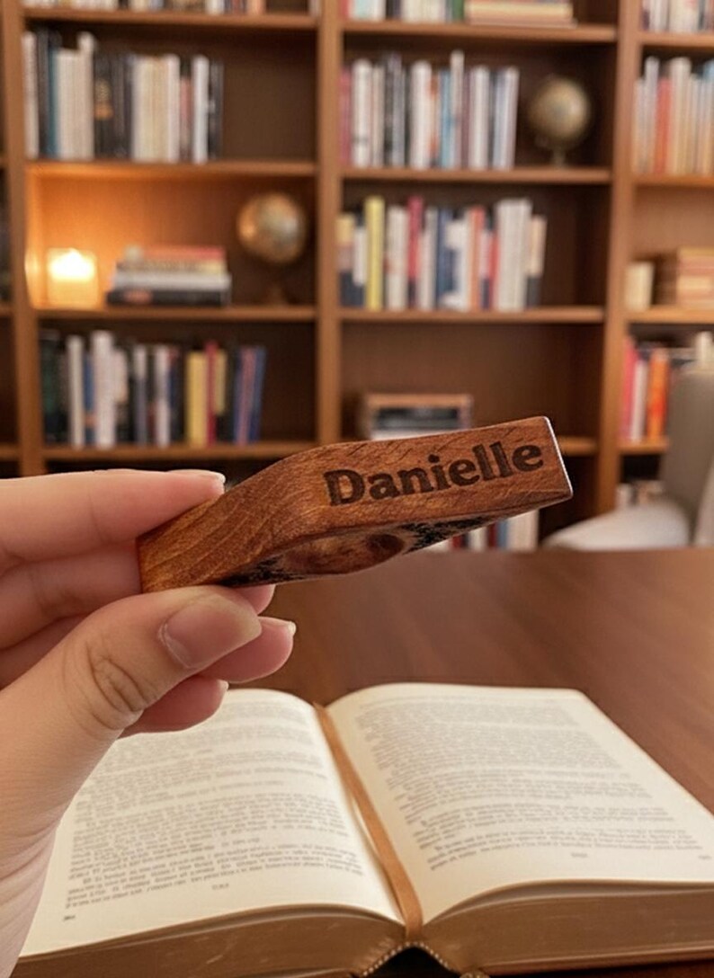 May include: A wooden bookmark with the name "Danielle" engraved on it. The bookmark is held above an open book, with a bookshelf in the background. The bookmark is made of a light brown wood.