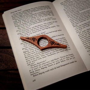 May include: A wooden book page holder with a floral design rests on an open book. The page holder is a warm brown color and has a circular opening for the finger. The book is open to page 216.