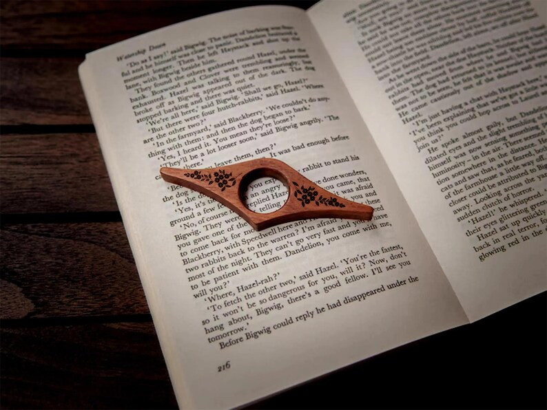 May include: A wooden book page holder with a floral design rests on an open book. The page holder is a warm brown color and has a circular opening for the finger. The book is open to page 216.