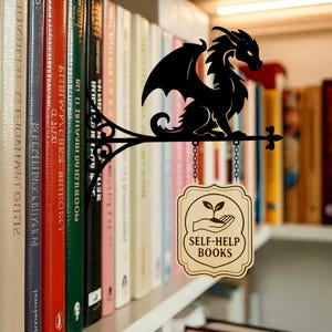 May include: A black metal dragon-shaped sign with a decorative flourish, hanging over a bookshelf. The sign has a wooden plaque that reads "SELF-HELP BOOKS" with a hand holding a plant. The sign is attached to a chain.