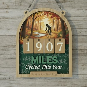 May include: Wooden cycling log with a scenic autumn forest illustration. The log displays "1907 MILES Cycled This Year" and "David's Cycling Log". The frame is light brown, with a chain for hanging.
