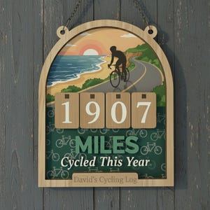 May include: Wooden cycling log with a beach scene and a cyclist on a road. The log displays "1907 MILES Cycled This Year" and "David's Cycling Log". The frame is light brown, with a chain for hanging.