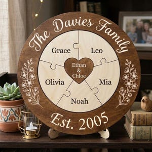 May include: A round wooden family puzzle with the text "The Davies Family" and "Est. 2005". The puzzle pieces are engraved with names: Grace, Olivia, Noah, Leo, and Mia, with a heart-shaped piece in the center with the names Ethan & Chloe.