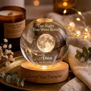 Puede incluir: Una esfera de cristal transparente con un diseño de luna llena, el texto "The Night You Were Born", el nombre "Anna" y la fecha "06.03.2025". La esfera se encuentra sobre una base de madera con la inscripción "Love You Forever".