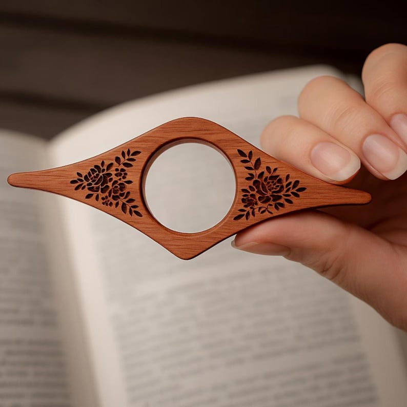 May include: A wooden book page holder with a diamond shape and a central circular opening. The holder is a warm brown color and features floral designs etched on each side of the opening. The holder is designed to keep a book open.