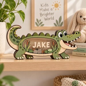 Könnte beinhalten: Eine hölzerne Spardose in Form eines grünen Krokodils mit einer transparenten Acrylfront. Der Name JAKE ist in weißen Buchstaben im Inneren des Körpers aufgedruckt, der verschiedene Metallmünzen enthält. Das Krokodil hat ein lächelndes Gesicht.