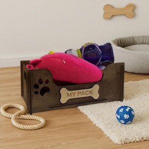 May include: A dark brown wooden pet toy box with a paw print cutout and a bone-shaped plaque that reads "MY PACK." The box is filled with dog toys, including a pink plush toy, a blue ball, and a rope toy. A wooden bone decorates the wall.