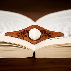 May include: A wooden book page holder with a floral design, resting on an open book. The holder is a warm brown color with a circular opening in the center. The floral design is etched into the wood.