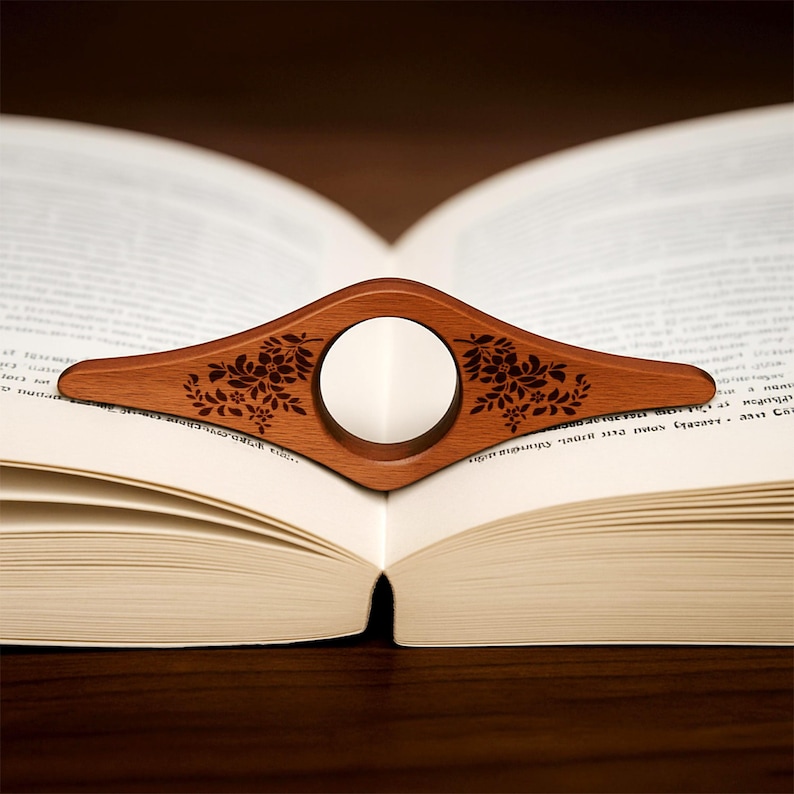 May include: A wooden book page holder with a floral design, resting on an open book. The holder is a warm brown color with a circular opening in the center. The floral design is etched into the wood.
