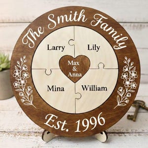 Puede incluir: Un rompecabezas familiar de madera personalizado con el texto "The Smith Family" y "Est. 1996". El rompecabezas presenta cuatro piezas con nombres y un corazón central. Acentos florales y un soporte completan el diseño.
