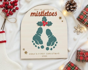 May include: A wooden Christmas sign with the text "MEET ME UNDER THE mistletoes" and baby footprints. The sign also includes the text "ANNA'S FIRST CHRISTMAS 2025". The sign is surrounded by Christmas decorations, including gifts and a stocking.