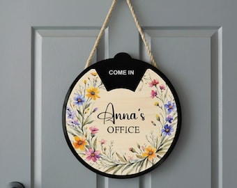 May include: Round wooden office sign with a black border and a floral design. The sign says "Anna's OFFICE" with the word "COME IN" at the top. The sign is hanging on a gray door with a black doorknob.