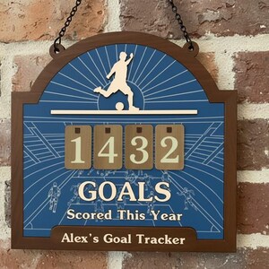 May include: Wooden soccer goal tracker with a blue background and the text "GOALS Scored This Year." The tracker displays the number 1432. The top features a soccer player kicking a ball. The bottom reads "Alex's Goal Tracker."