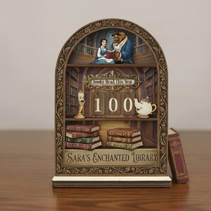 Può includere: Un elemento decorativo a forma di libreria in legno con personaggi de La Bella e la Bestia. Il design include uno scaffale con pile di libri, il testo "Books Read This Year" e "Sara's Enchanted Library", e il numero "100".