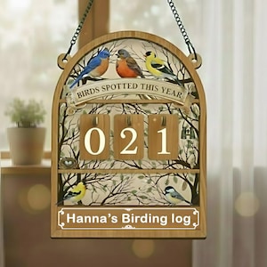 Könnte beinhalten: Holzschild für Vogelbeobachtung mit dem Text "Birds Spotted This Year" und "Hanna's Birding log". Das Schild zeigt Illustrationen von Vögeln und einen Zähler mit der Anzeige "021". Das Schild hängt an einer Kette.