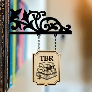 May include: Black metal bookshelf bracket with a decorative floral design. A wooden sign hangs from chains, displaying "TBR" above a stack of books. The bracket is mounted on a shelf with a row of colourful books.
