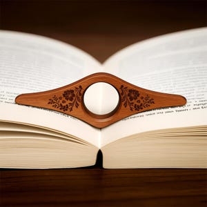May include: A wooden book page holder with a floral design, resting on an open book. The holder is a warm brown color and features a circular opening in the center. The floral pattern is etched into the wood.