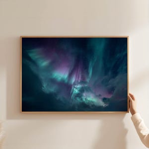 Könnte beinhalten: Ein gerahmter Druck mit einer lebendigen Darstellung der Aurora Borealis. Das Kunstwerk zeigt wirbelnde Bänder in Türkis, Lila und Dunkelblau, die ein Gefühl des Staunens und der Schönheit des Nachthimmels hervorrufen. Der Druck ist in einem hellbraunen Holzrahmen eingefasst.