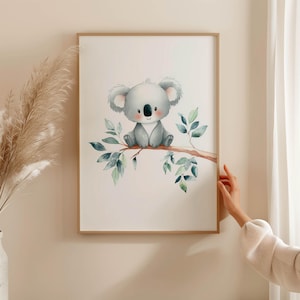 Könnte beinhalten: Ein gerahmter Aquarelldruck mit einem niedlichen Koala-Bären, der auf einem Ast mit grünen Blättern sitzt. Der Koala ist grau mit rosa Nase und Ohren. Das Kunstwerk hat einen hellbeigen Hintergrund und einen Holzrahmen.