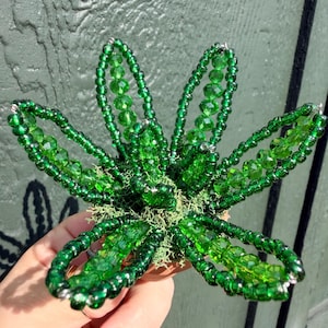 Pode incluir: Uma escultura floral decorativa feita de contas de vidro verde vibrante. A pe&ccedil;a apresenta cinco p&eacute;talas distintas constru&iacute;das com pequenas contas de sementes e contas de cristal facetadas maiores, centradas numa base de musgo verde claro.