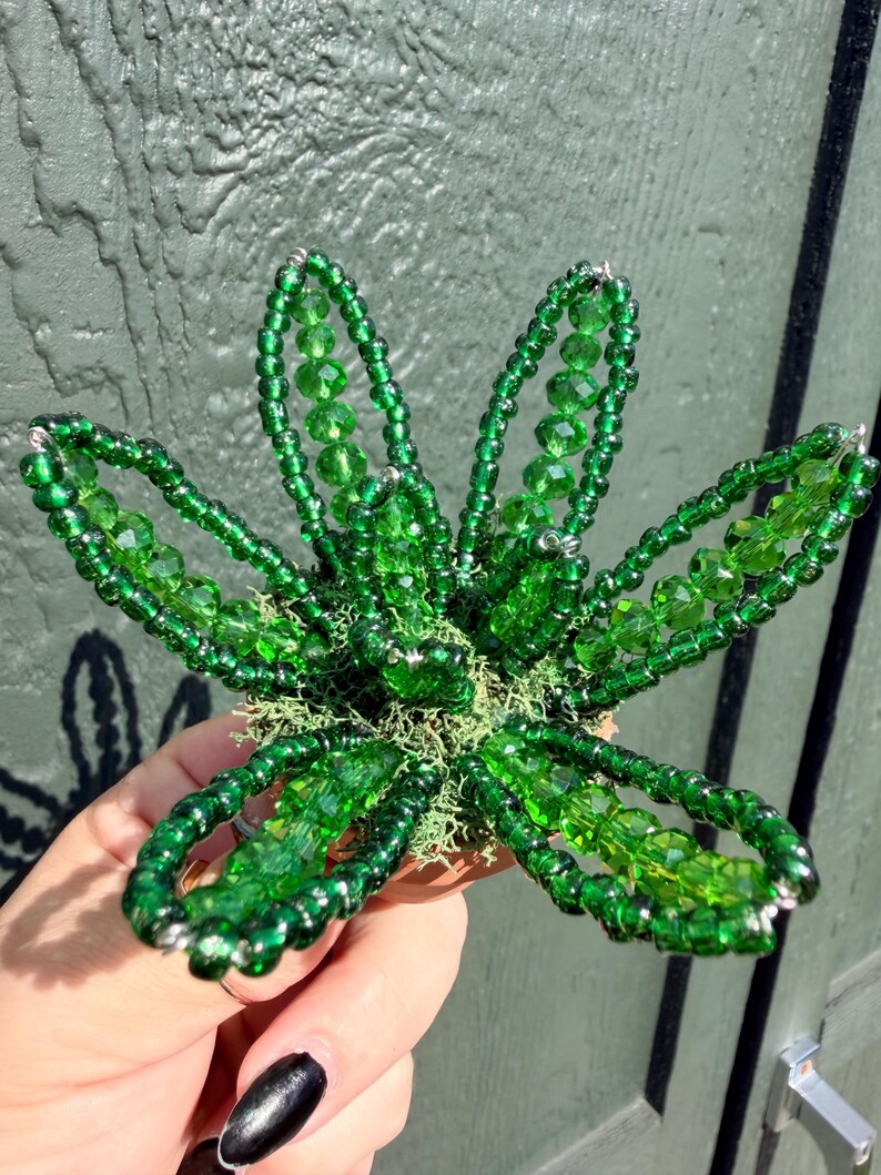 Pode incluir: Uma escultura floral decorativa feita de contas de vidro verde vibrante. A pe&ccedil;a apresenta cinco p&eacute;talas distintas constru&iacute;das com pequenas contas de sementes e contas de cristal facetadas maiores, centradas numa base de musgo verde claro.