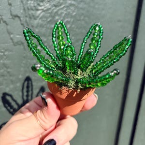 Pode incluir: Uma pequena planta suculenta decorativa feita de contas de vidro verde transl&uacute;cido, com folhas pontiagudas dispostas num padr&atilde;o circular. A planta de contas est&aacute; colocada num vaso de barro cor de terracota cheio de musgo verde.