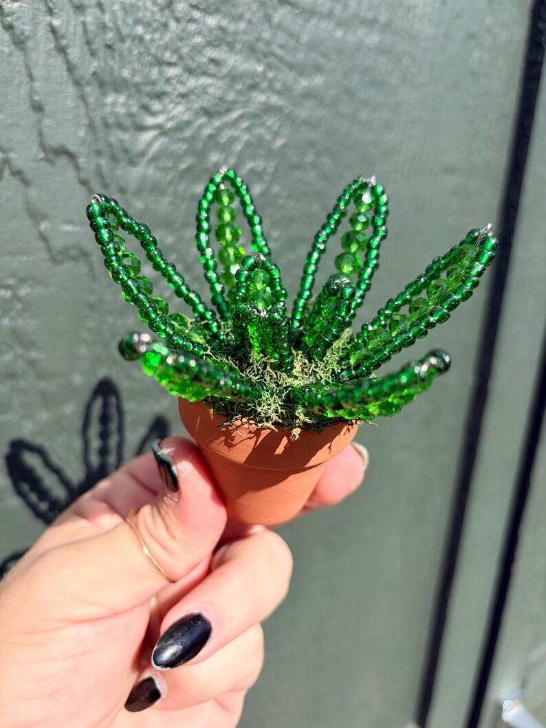 Pode incluir: Uma pequena planta suculenta decorativa feita de contas de vidro verde transl&uacute;cido, com folhas pontiagudas dispostas num padr&atilde;o circular. A planta de contas est&aacute; colocada num vaso de barro cor de terracota cheio de musgo verde.
