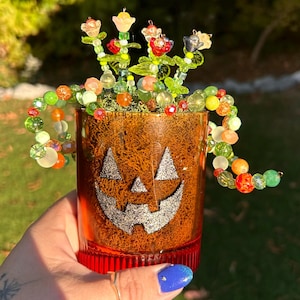 May include: A decorative Halloween-themed glass with a jack-o'-lantern face design. The orange glass is filled with decorative beads and faux flowers. A beaded garland of colorful beads surrounds the top of the glass.