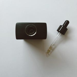 May include: A black bottle with a dropper. The bottle is rectangular with a circular opening. The dropper has a black top and a clear glass tube with a yellow liquid inside. The items are on a white surface.