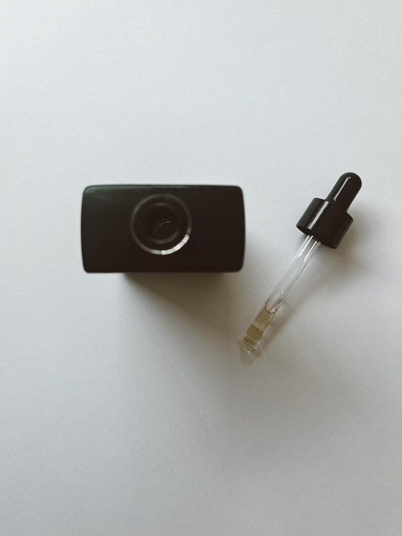 May include: A black bottle with a dropper. The bottle is rectangular with a circular opening. The dropper has a black top and a clear glass tube with a yellow liquid inside. The items are on a white surface.
