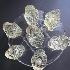 Op de afbeelding: Zeven heldere, gebeeldhouwde glazen objecten, mogelijk decoratief of functioneel, zijn gerangschikt op een heldere, cirkelvormige plaat. De objecten hebben een afgeronde vorm met ingewikkelde, getextureerde details en een lichte gele tint.