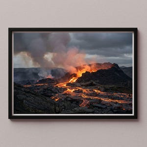 May include: A framed photograph captures a volcanic eruption. Molten lava flows down the mountain, glowing orange against the dark, rocky terrain. Smoke billows into a cloudy, gray sky, creating a dramatic landscape scene.