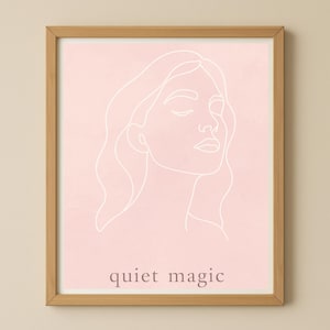 May include: A framed art print featuring a minimalist line drawing of a woman's face with closed eyes and flowing hair. The artwork is in white on a soft pink background, with the words "quiet magic" at the bottom.