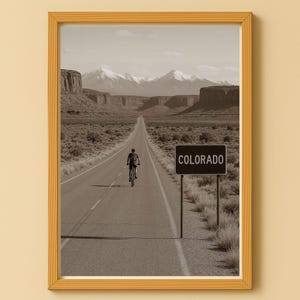 Könnte beinhalten: Sepiafarbener gerahmter Druck mit einem Radfahrer auf einer langen Straße, die zu Bergen führt. Ein Schild mit der Aufschrift "COLORADO" befindet sich auf der rechten Seite. Das Bild vermittelt Abenteuerlust und Reise.