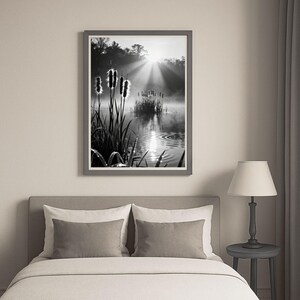 May include: Monochrome framed print depicting a tranquil lake scene. The image features bulrushes and other aquatic plants, with sunlight piercing through the mist. The artwork is displayed above a bed with grey pillows and a lamp.