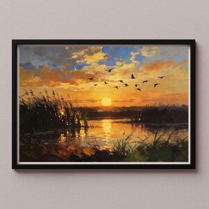 Op de afbeelding: Een ingelijst landschapsschilderij toont een zonsondergang boven een moeras. De lucht is een mix van oranje, gele en blauwe tinten, met silhouetten van vogels in vlucht. Riet en grassen omlijsten de waterkant en reflecteren het warme licht.
