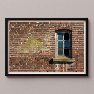 May include: A framed photograph of a brick wall with a weathered window. The wall is made of red bricks, with some green discoloration. The window has a wooden frame and broken glass, with a view of the sky.