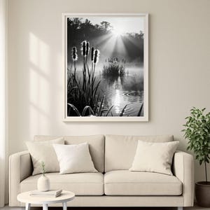 May include: Black and white framed photograph of a tranquil pond scene. Reeds and cattails are in the foreground, with a misty pond reflecting the sun's rays. The photograph is displayed above a beige sofa with white cushions.