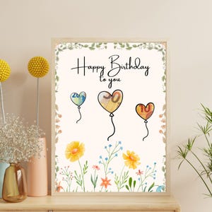 Könnte beinhalten: Ein gerahmter Druck mit den Worten "Happy Birthday to you" in eleganter Schrift. Das Kunstwerk zeigt drei herzförmige Ballons, jeder mit einer Zahl, und einen floralen Rand. Die Ballons sind farblich so gestaltet, dass sie an Währung erinnern.