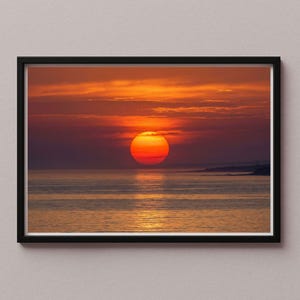 Pode incluir: Fotografia emoldurada de um pôr do sol sobre o oceano. O sol, uma grande esfera laranja, está perto do horizonte, refletindo-se na água. O céu é uma mistura de tons de laranja, vermelho e roxo, com algumas nuvens visíveis.
