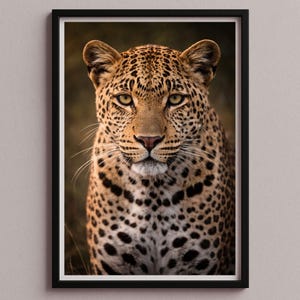 May include: A framed print featuring a close-up portrait of a leopard. The leopard has a tan coat with black spots, yellow eyes, and a pink nose. The print is in a black frame and is set against a neutral background.