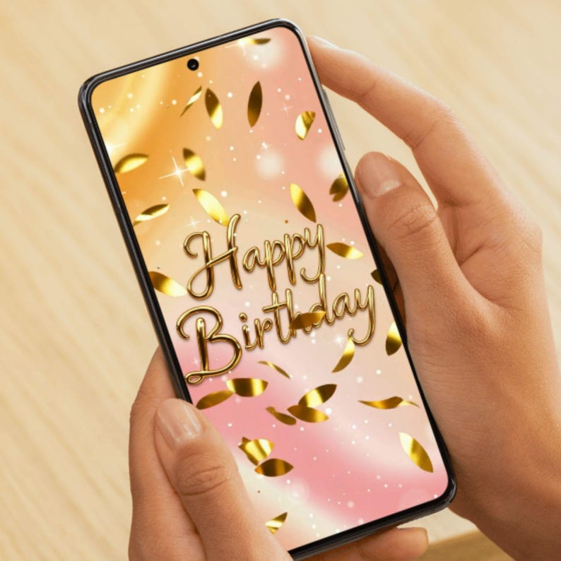 May include: A smartphone displays a birthday greeting. The screen shows the words "Happy Birthday" in gold script, surrounded by gold confetti against a gradient background of pink and orange. The phone is held in two hands.